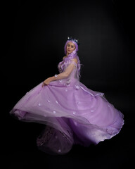 Fototapeta premium Full length portrait of girl wearing long purple fantasy ball gown with crown and pink hair, standing pose with elegant gestural movements against a studio background.