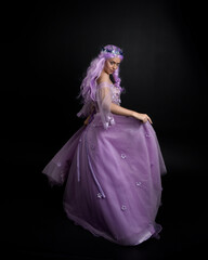 Full length portrait of girl wearing long purple fantasy ball gown with crown and pink hair, standing pose with back to the camera  against a studio background.