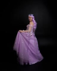 Full length portrait of girl wearing long purple fantasy ball gown with crown and pink hair, standing pose with back to the camera  against a studio background.