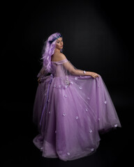 Obraz na płótnie Canvas Full length portrait of girl wearing long purple fantasy ball gown with crown and pink hair, standing pose with back to the camera against a studio background.