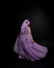 Full length portrait of girl wearing long purple fantasy ball gown with crown and pink hair, standing pose with back to the camera  against a studio background.