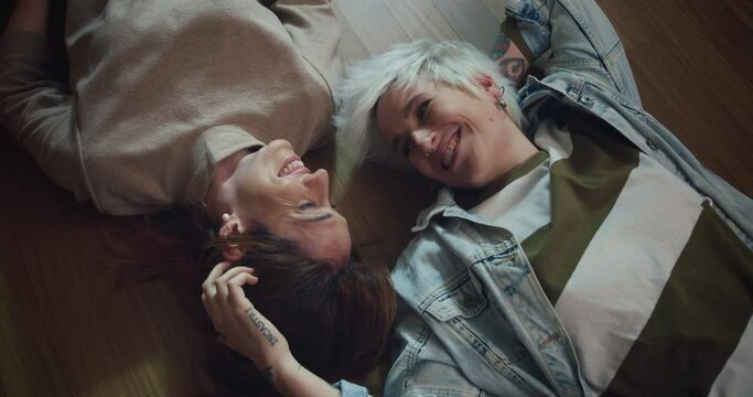 Cinematic shot of carefree happy homosexual female gay and transgender man couple in love is enjoying time together is speaking and caressing each other with affection while lying on floor at home.