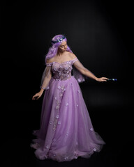 Fototapeta premium Full length portrait of girl wearing long purple fantasy ball gown with crown and pink hair, standing pose holding a wand against a studio background.