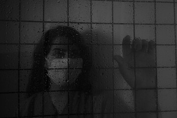 selective focus of  nurse with face mask behind bars, looking out of wet window. she is holding fence.