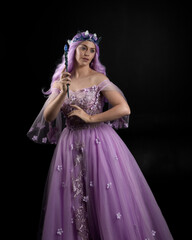 Close up portrait of girl wearing long purple fantasy ball gown with butterfly crown and pink hair, against a  dark studio background.
