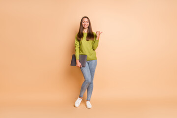 Fototapeta na wymiar Photo of lady hold netbook indicate finger empty space wear green pullover jeans footwear isolated beige color background