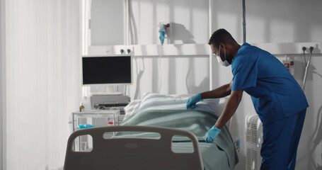 Expert young male nurse in mask working at hospital and making bed - Powered by Adobe