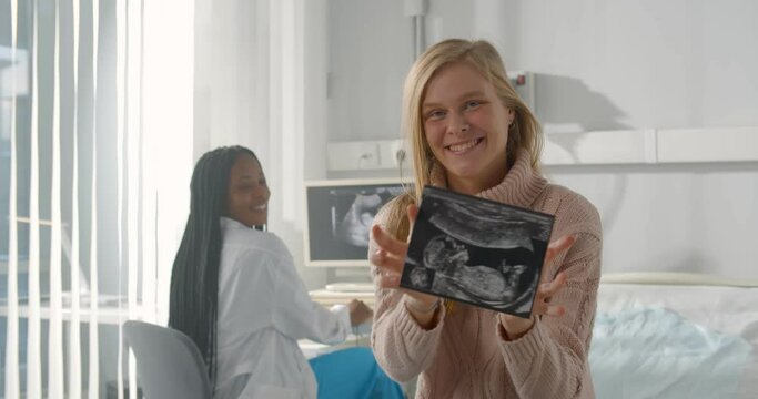 Smiling pregnant woman holding ultrasound baby image sitting in gynecologist office