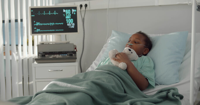 Afro-american kid lying in hospital bed with oxygen tube and plush toy