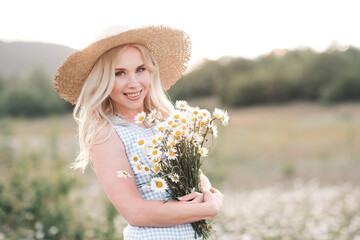 Smiling beautiful blonde woman 25-29 year old holding bouquet of fresh daisy wearing straw hat and...