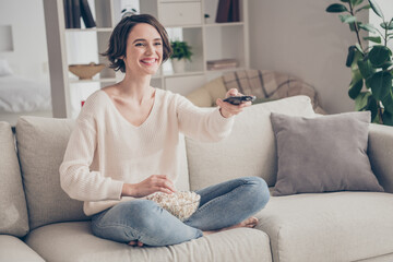 Obraz premium Full length photo of positive charming girl sit on couch hold remote switch channels toothy smile have fun home indoors