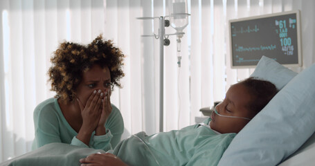 Upset young african woman crying sitting near little daughter sleeping in bed at hospital