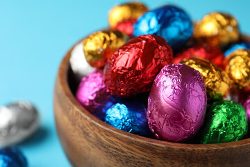 Fototapeta na wymiar Wooden bowl with chocolate eggs wrapped in colorful foil on light blue background, closeup