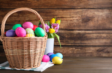 Colorful Easter eggs in wicker basket and tulips on wooden background. Space for text