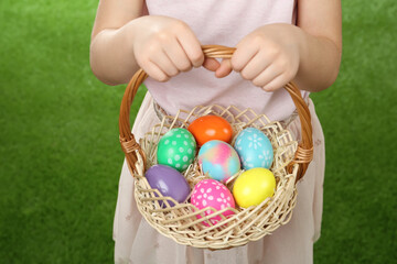 Fototapeta na wymiar Little girl with basket full of Easter eggs on green grass, closeup