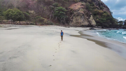 A man walking on an idyllic Koka Beach. Hidden gem of Flores, Indonesia. He is enjoying a solo escape. Waves gently washing the shore. There are hills in the back. Happiness, adventure and discovering