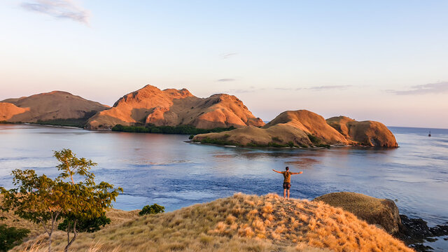 A man standing on top of a small island, enjoying the morning sun over Komodo National Park, Flores, Indonesia. Golden hour over the islands and sea. Some boats anchored to the bay. New day beginning