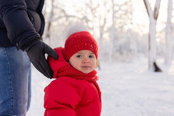 Fototapeta na wymiar Portrait of a little girl in a knitted hat and red overalls, she is standing in a snowy park, and her dad is holding her.