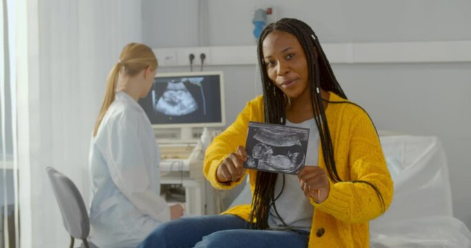 Happy pregnant black woman showing sonogram picture at camera sitting in ultrasound office