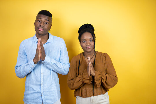 Young African American Couple Standing Over Yellow Background Begging And Praying With Hands Together With Hope Expression On Face Very Emotional And Worried