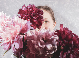 Large blurred bouquet of pink peonies covering face young woman on grey background. Focus are on one her eye.