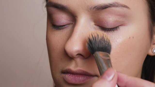 The makeup artist applies a beige liquid foundation on the female face with a brush. Close-up of smokey eyes of a young woman