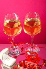 Two glasses of champagne tied with ribbons on a pink background next to a ring in a box and a candle in a candlestick.