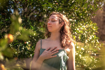 Portrait of young woman in nature with green dress. Touching her chest with eyes closed. Sunlight