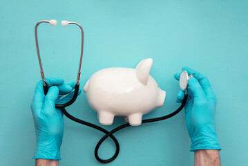 Health care cost. Doctor in surgical gloves holding a white piggy bank and stethoscope