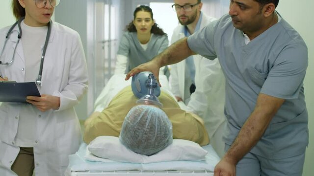 Tilt up shot of team of paramedics giving ventilation to emergency patient with BVM and pushing stretchers towards the camera through hospital corridor
