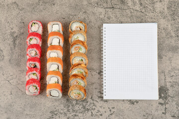 Assortment of tasty sushi rolls and blank notebook on marble background