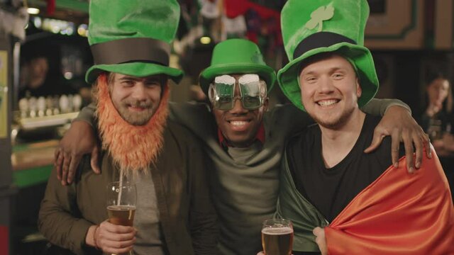 Slow-motion medium portrait of cheerful multi-ethnic men in funny green hats with clover posters celebrating St Patricks Day in pub, drinking beer and having fun together