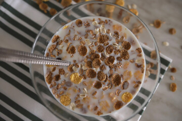 A cup of muesli is on a stylish white and green napkin. Flakes are scattered on the table. Muesli is covered with milk and a spoon lies in a cup with muesli.