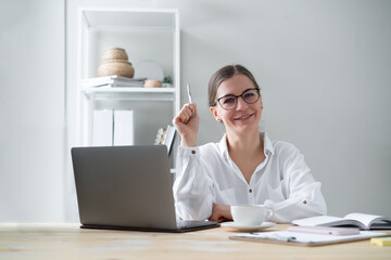 Happy young woman psychologist sits table with laptop and holds  pen in her raised hand. She looks camera smiling.