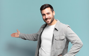 handsome adult blond man feeling happy and cheerful, smiling and welcoming you, inviting you in...