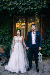 The newlyweds in love stand against the background of a wall, a growing green bush and a window. Wedding portrait of a groom in a suit and a beautiful bride.