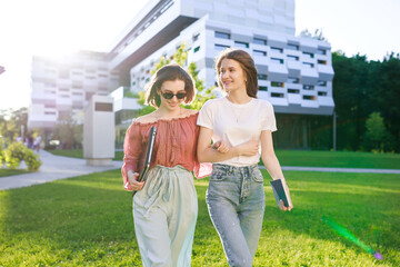 Two friends from college are walking down the street after school in a T-shirt, jeans and sunglasses, holding a computer and books in their hands, talking.