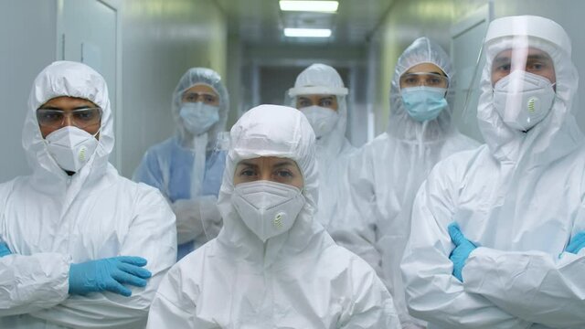 Team of multiethnic male and female doctors in protective suits, face shields, masks and gloves standing together in hospital and posing for camera during workday in time of coronavirus pandemic