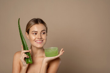 Smiling happy young female model holding aloe leaf and jar of aloe gel on beige background. The...