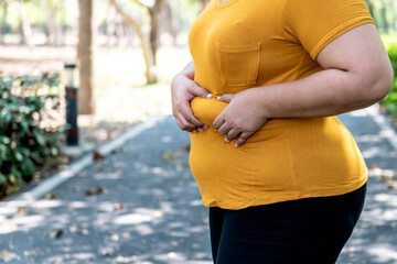 Fat women  Using two hands to hold the excess fat In the area of her waist, with blur park background to obesity and health care concept. - obrazy, fototapety, plakaty
