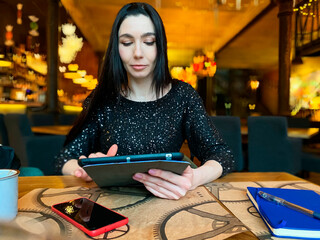 A girl with dark hair is working in a cafe with a tablet, a phone and a notebook are nearby....