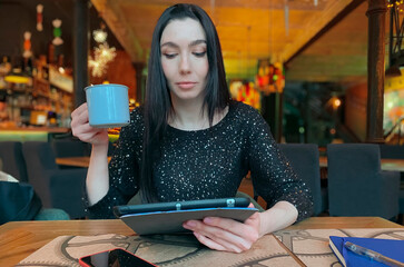 A girl with dark hair is working in a cafe with a tablet, a phone and a notebook are nearby....