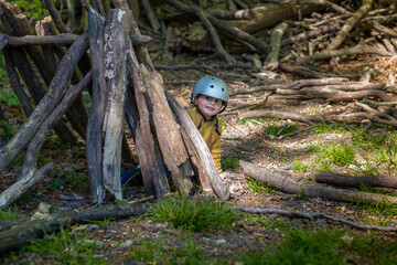 A young boy is playing in the forest in summer or spring. Toddler is building a  wooden hut of logs and branches. Child hiding in wigwam created  in the park.