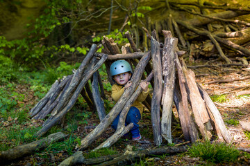 A young boy is playing in the forest in summer or spring. Toddler is building a  wooden hut of logs and branches. Child hiding in wigwam created  in the park.