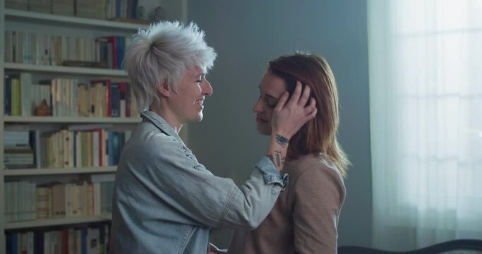 Cinematic shot of carefree happy homosexual female gay and transgender man couple in love is enjoying time together is hugging and kissing with affection as sign of timeless forever love at home.