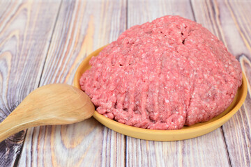Minced meat in a wooden plate. A wooden spoon lies nearby. The plate stands on a wooden table.