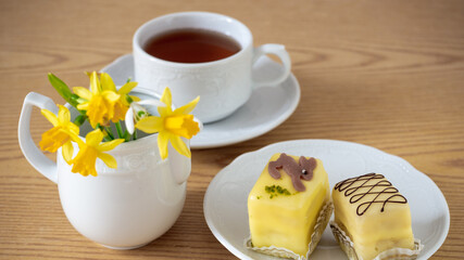 Easter breakfast, afternoon tea yellow marzipan glazed cakes, petit fours decorated with a bunny, white porcelain cup of tea. Daffodil flowers in a saucer on a wooden table. Close up, macro