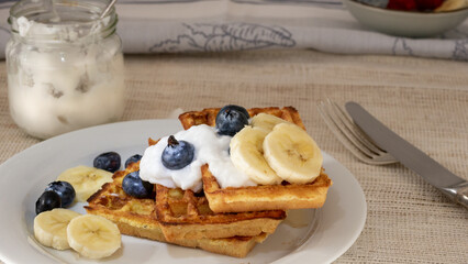 Stack of freshly made waffles with coconut yogurt, banana and blueberry on a place and fork for Sunday breakfast.