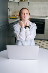 Portrait of tired redhead young business woman wit closed eyes having neck pain during working at laptop computer while sitting at desk in home workplace. Concept of remote working from home office.