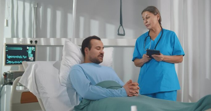 Female doctor discussing medical report with male patient on digital tablet in ward at hospital.